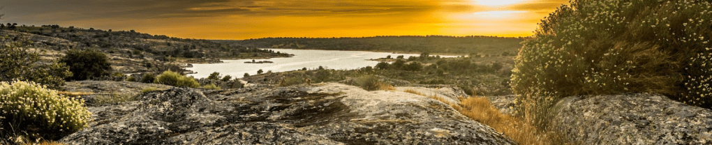 Paisaje natural con rocas grandes, vegetación, y un lago bajo un atardecer dorado.