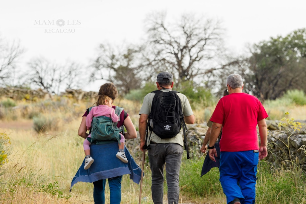 Cuatro personas caminando por un sendero natural, una de ellas lleva a un niño en una mochila. El grupo está rodeado de vegetación y árboles.