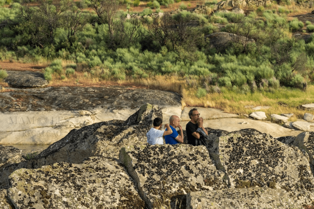 Tres personas sentadas en unas rocas, observando el paisaje natural con vegetación al fondo.