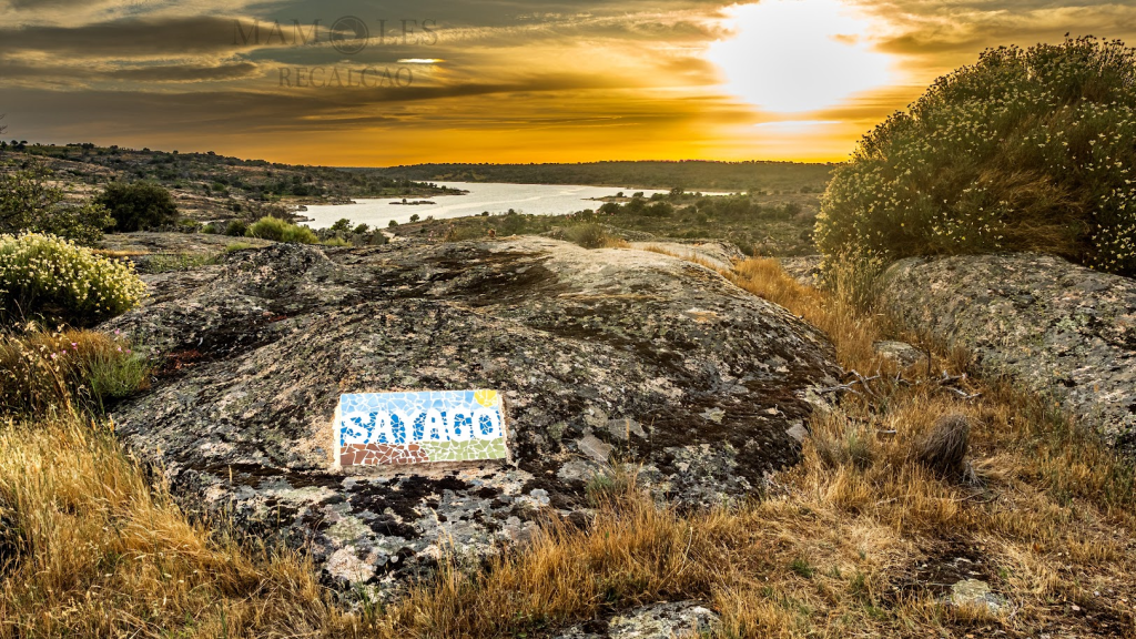 Vista del atardecer sobre un lago con un letrero colorido que dice 'Santiago' en un paisaje rocoso.