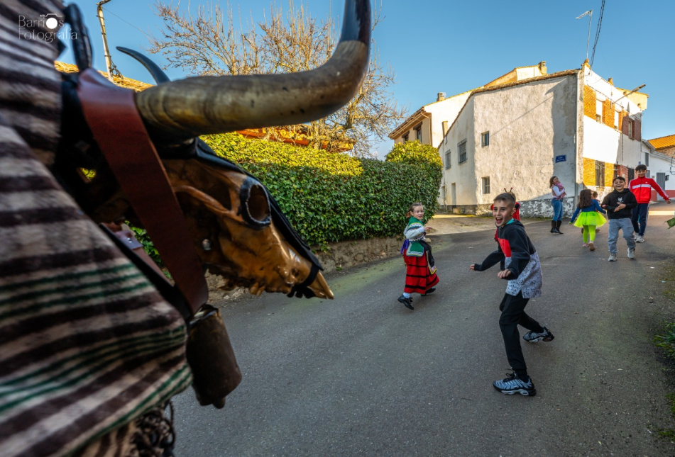 Niños corriendo y riendo en una calle, mientras un hombre disfrazado con una máscara de toro se acerca, rodeados de casas y vegetación.