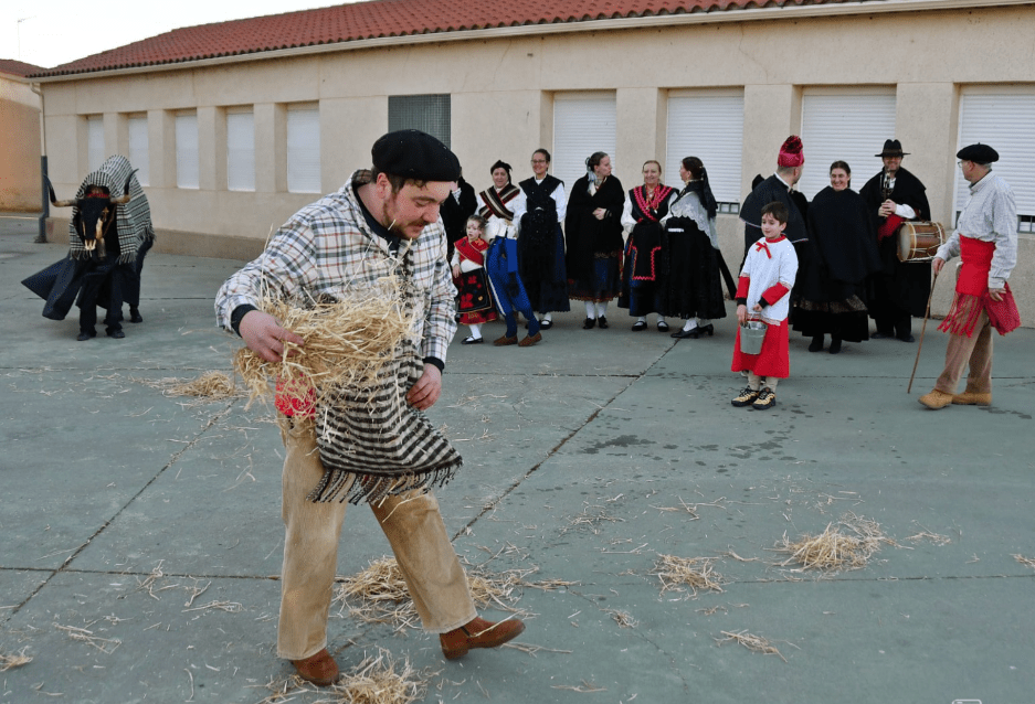 Hombre en ropa tradicional lanzando paja en un evento festivo, con un grupo de personas vestidas con trajes típicos al fondo.