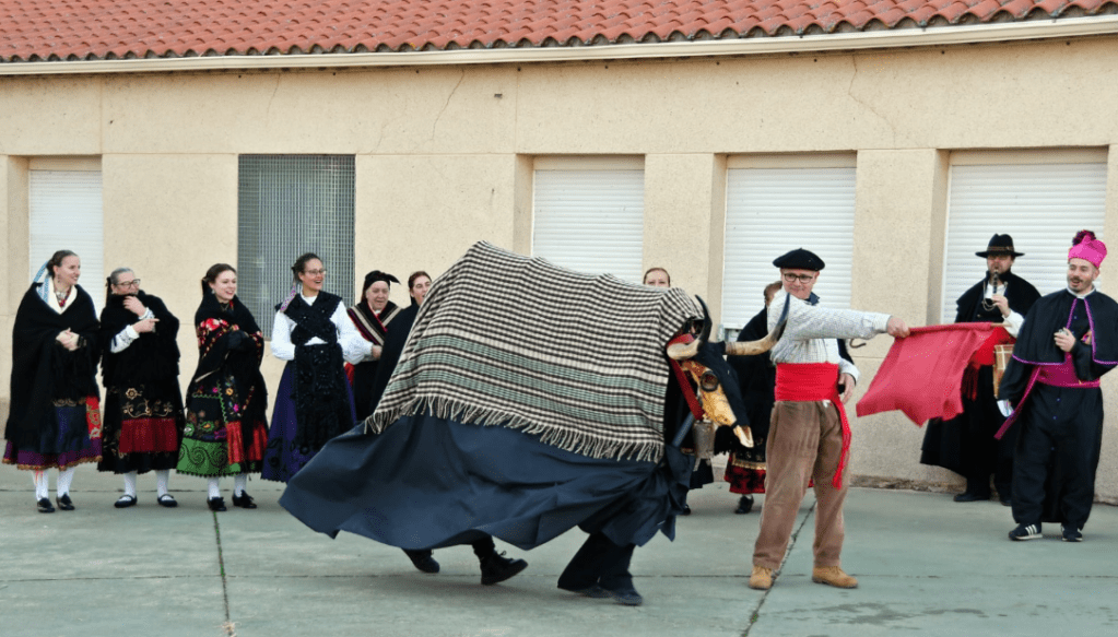 Grupo de personas bailando en trajes tradicionales, uno de ellos sostiene una manta mientras otro lleva un pañuelo rojo.