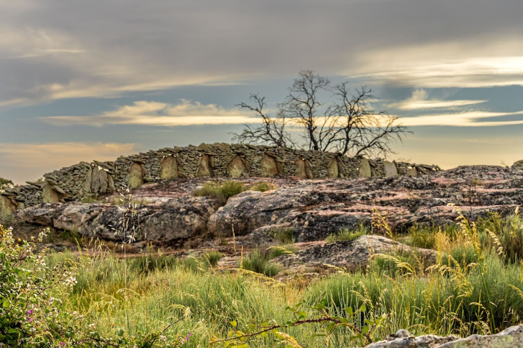 Un paisaje con formaciones rocosas cubiertas de hierba y un antiguo círculo de piedras en el fondo, bajo un cielo nublado.