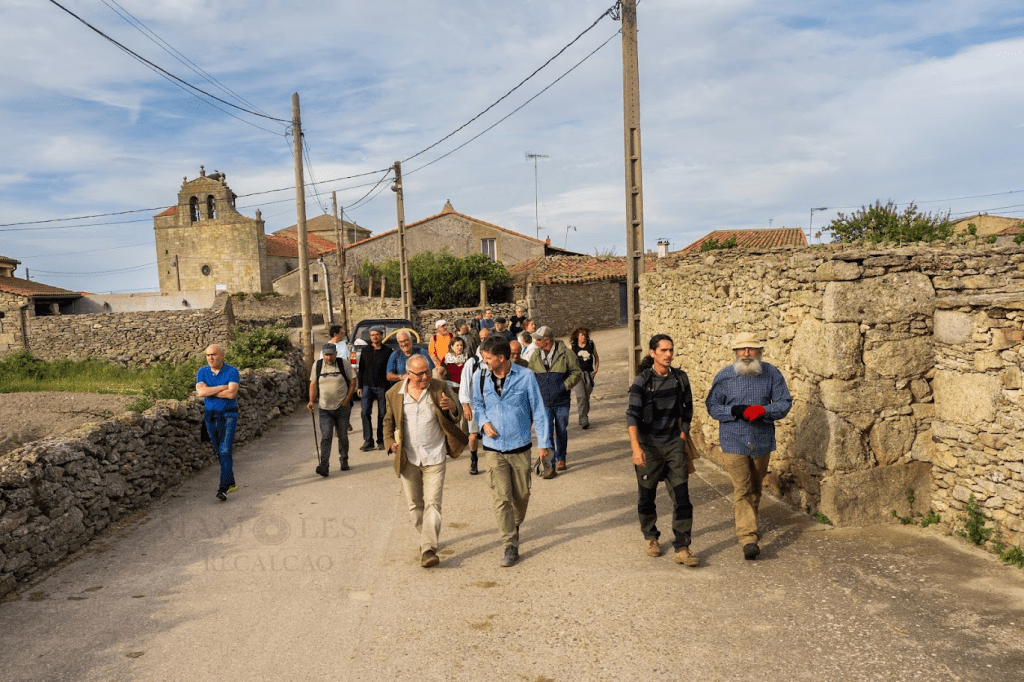 Grupo de personas caminando por un camino rural, con casas de piedra y un campanario al fondo bajo un cielo parcialmente nublado.